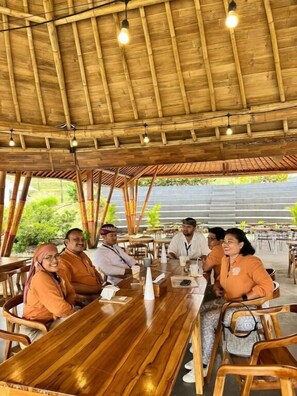 A group of people seated at a wooden table under a bamboo roofed area with hanging lights. - The Blooms Villa (Baturiti)