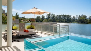 Uma área moderna de piscina ao ar livre com estrutura de vidro, uma espreguiçadeira, um guarda-sol e vista para um lago.