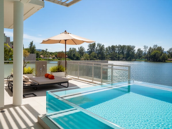 A modern outdoor pool area with a glass enclosure, a lounge chair, an umbrella, and a view of a lake.