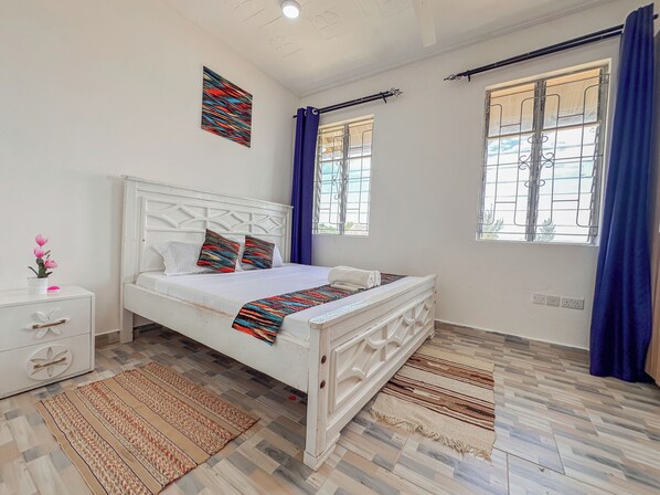 Un dormitorio con cama, mesita de noche, ventana con cortinas y una lámpara fijada a la pared.