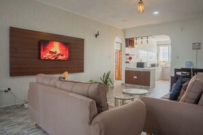 Una sala de estar con sofás, un televisor montado en la pared, una mesita y una cocina al fondo.