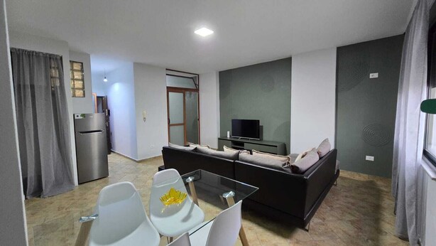 A modern living room with a sofa, a glass coffee table, and a TV. There is a kitchen area with a refrigerator and a dining table with chairs. - Tirent Apartment-One Bedroom Central (Tirana)