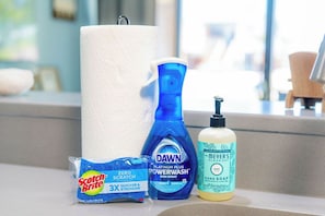 Un plan de vasque de salle de bain avec un rouleau d'essuie-tout, une bouteille de liquide vaisselle Dawn bleu, une éponge à récurer Scotch-Brite « Zéro Rayures » et une bouteille de savon pour les mains Carex Clean Day pour hommes.