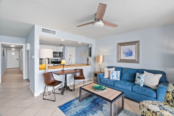 A modern living room with a blue sofa, a coffee table, and a dining area with chairs and a kitchen in the background.