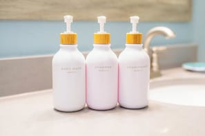 Tre flasker sæbe, shampoo og balsam på en badeværelseshylde.