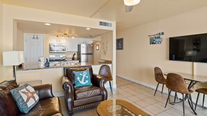 A living room with a brown leather sofa, a dining area with brown chairs, a kitchen in the background, and a ceiling fan. - Paradise | Waterfront Comfort at Laguna Reef 411 (Rockport)