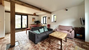 Ein Wohnzimmer mit Sofa, Sessel und Couchtisch. Es gibt einen Essbereich mit Tisch und Stühlen. Der Raum hat einen Ziegelboden und Holzbalken an der Decke.