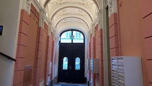 Un corridoio con decorazioni elaborate al soffitto, un portone doppio in fondo e un quadro elettrico a parete.