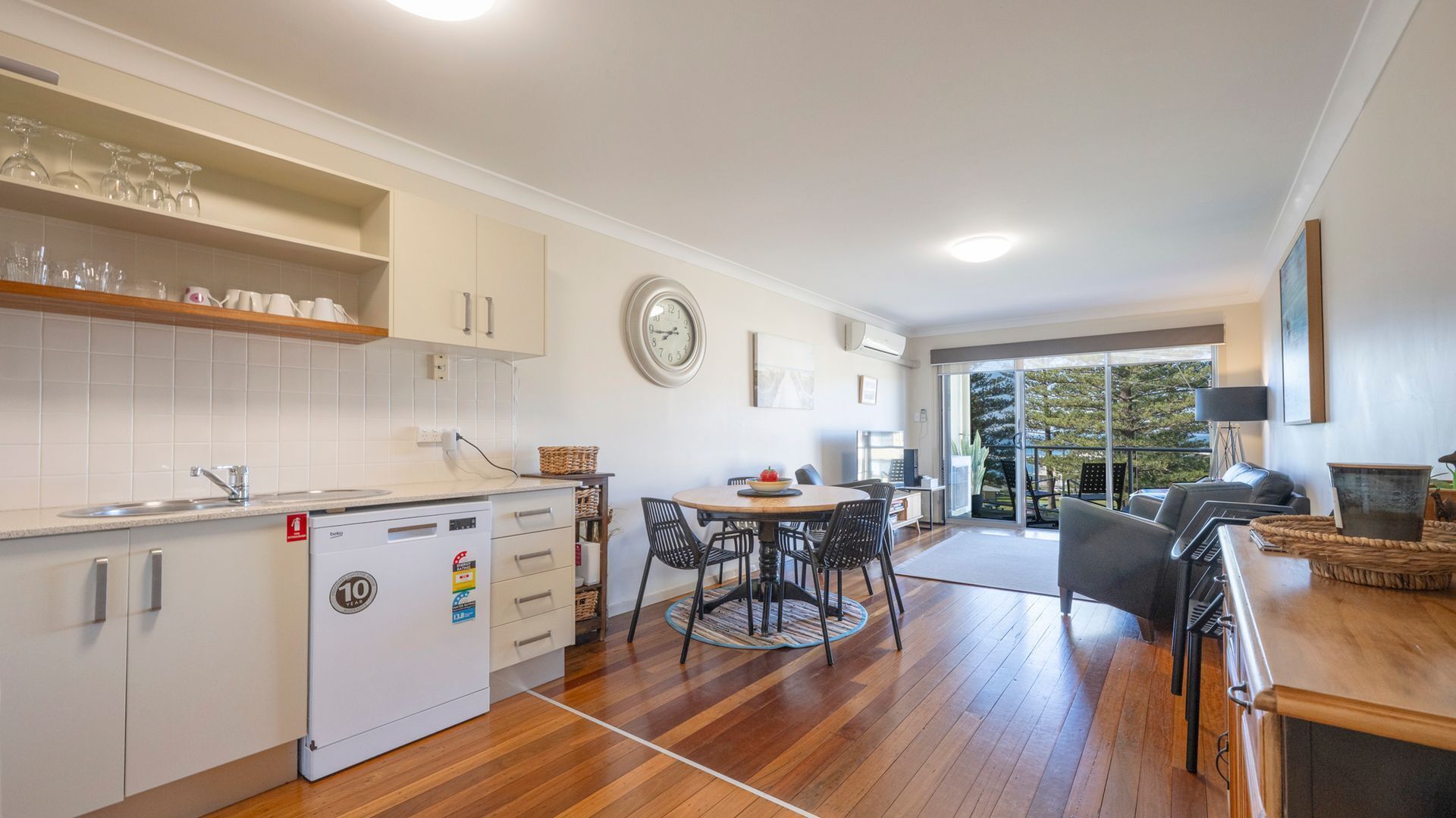 A modern kitchen with a built-in dishwasher, a dining area with a round table and chairs, and a view of the outdoors through a glass door.