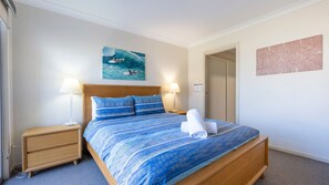 Quarto de hotel com uma cama, duas luminárias de cabeceira, uma mesa de apoio, um quadro na parede e uma porta que dá para outro cômodo.