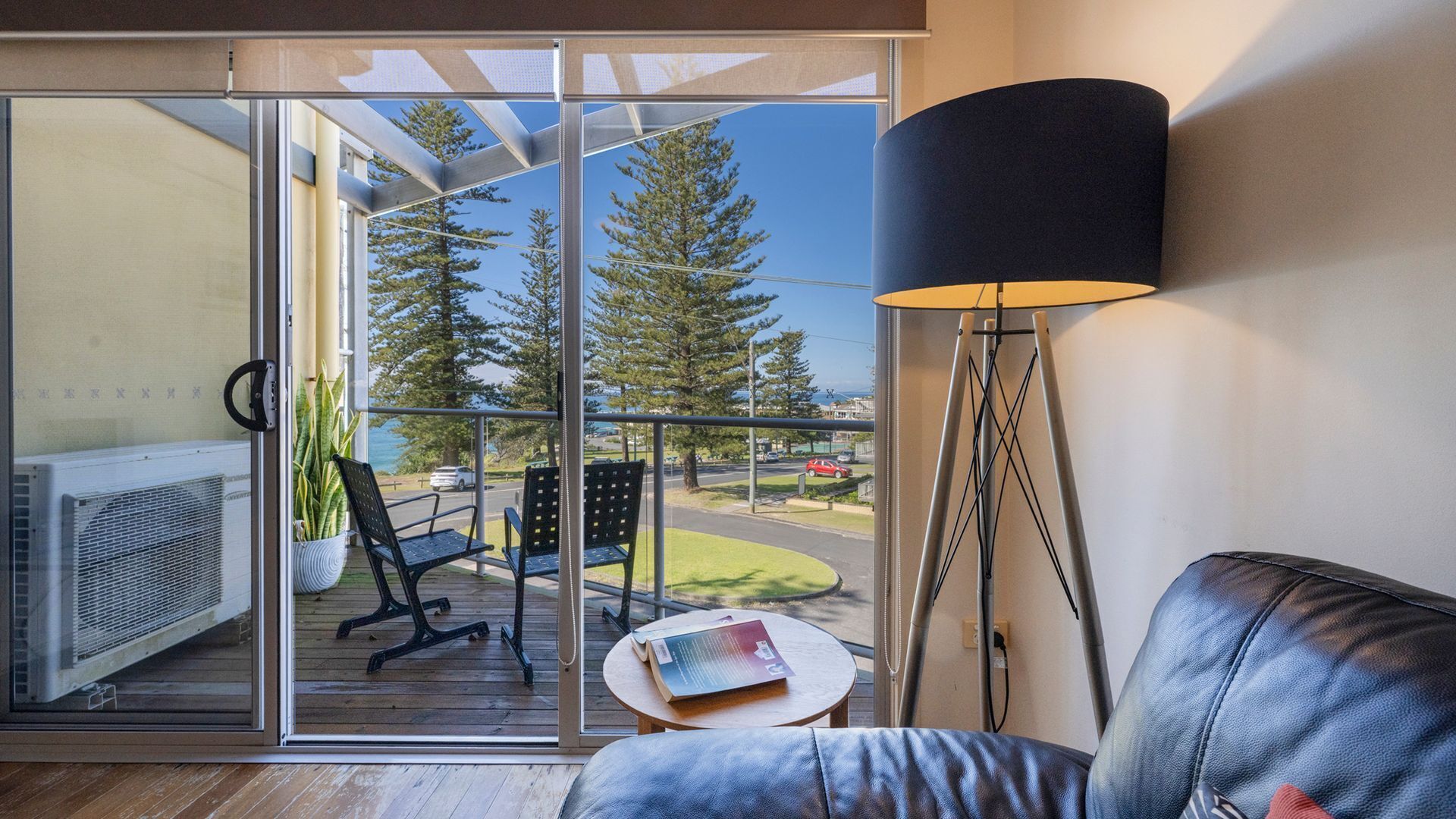 A modern living room with a sliding glass door leading to a balcony, a leather sofa, a floor lamp, and a small table with a book.