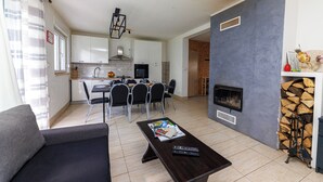 Een moderne woonkamer met een bank, een salontafel met tijdschriften en een open haard. Er is een eethoek met een tafel en stoelen, en een keuken op de achtergrond.