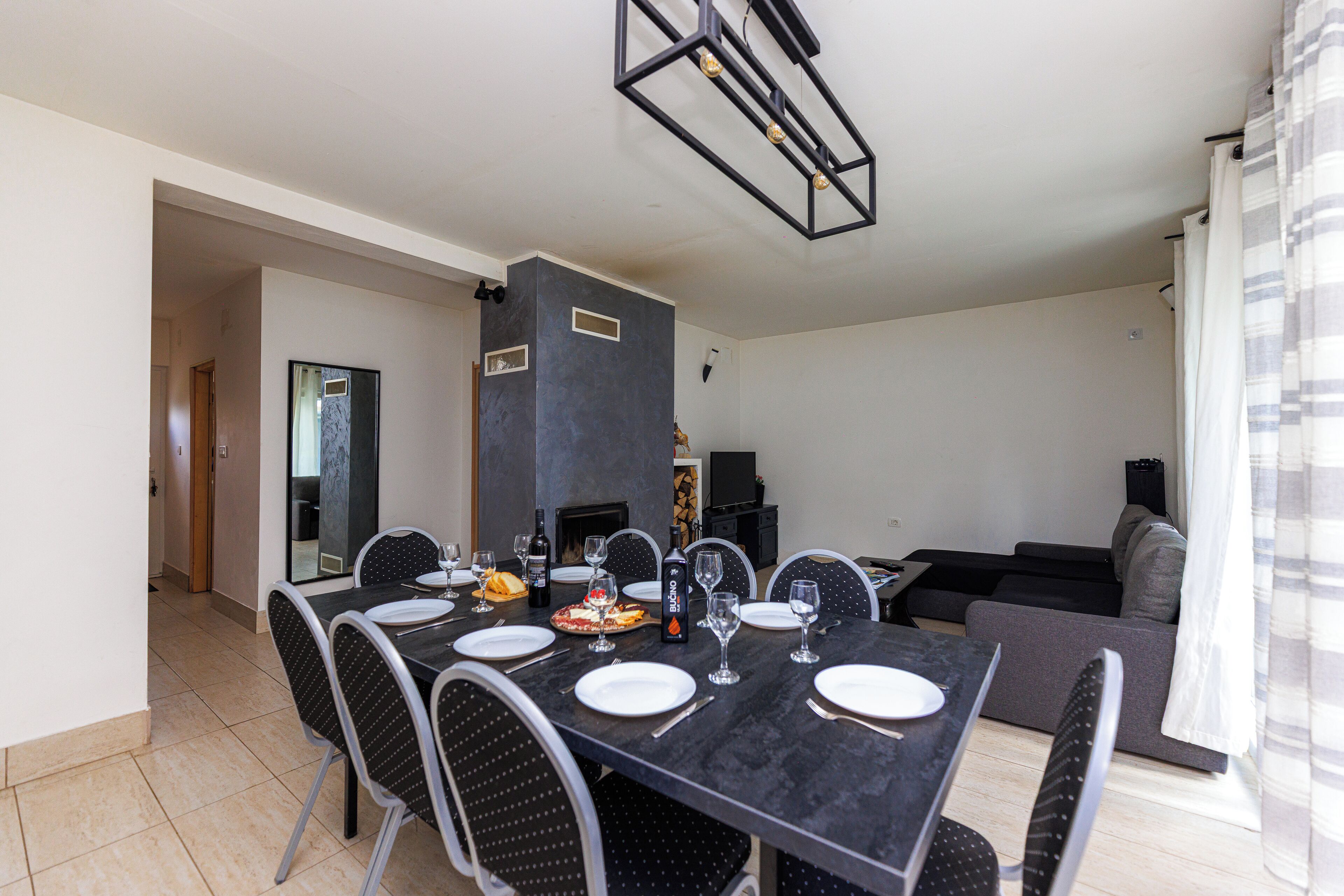 Un comedor con una mesa negra dispuesta para seis personas, rodeada de sillas. Se ve una chimenea al fondo, y un sofá está ubicado a la derecha.