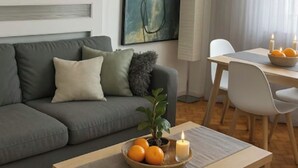 A modern living room with a grey sofa, a wooden coffee table with oranges and a candle, a dining area with a white table and chairs, and a large abstract painting on the wall. - A cocoon in Porto .4 to 6 person modular apartment, practical and convivial (Porto)