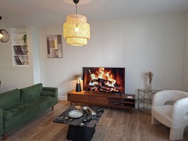 Una sala de estar con un sofá verde, una mesa de centro, un televisor que muestra una chimenea, una lámpara y una silla blanca.