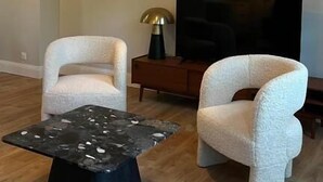 Un salon moderne avec deux fauteuils blancs, une table basse noire, un luminaire suspendu jaune et un téléviseur à écran plat posé sur un meuble en bois.