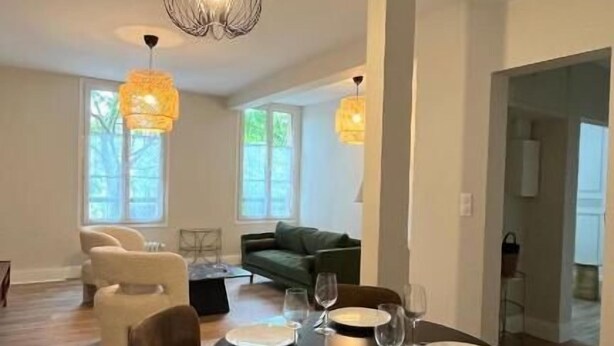 Un coin repas avec une table ronde dressée pour quatre personnes, entourée de chaises marron. Deux lampes suspendues sont placées au-dessus de la table, et un canapé vert est visible à l’arrière-plan.