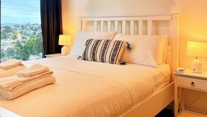 Una cama bien hecha con sábanas blancas, un cojín a rayas y vistas a un paisaje urbano a través de un gran ventanal.
