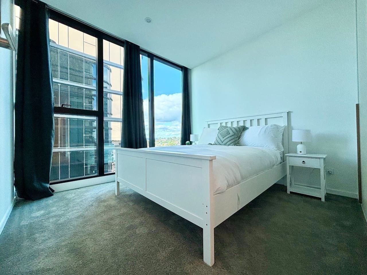 A bedroom with a bed, a nightstand, a window with curtains, and a view of buildings.
