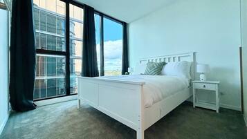 A bedroom with a bed, a nightstand, a window with curtains, and a view of buildings.