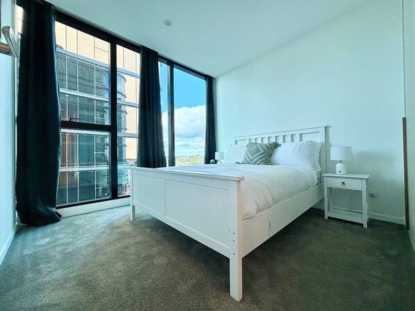 A bedroom with a bed, a nightstand, a window with curtains, and a view of buildings.