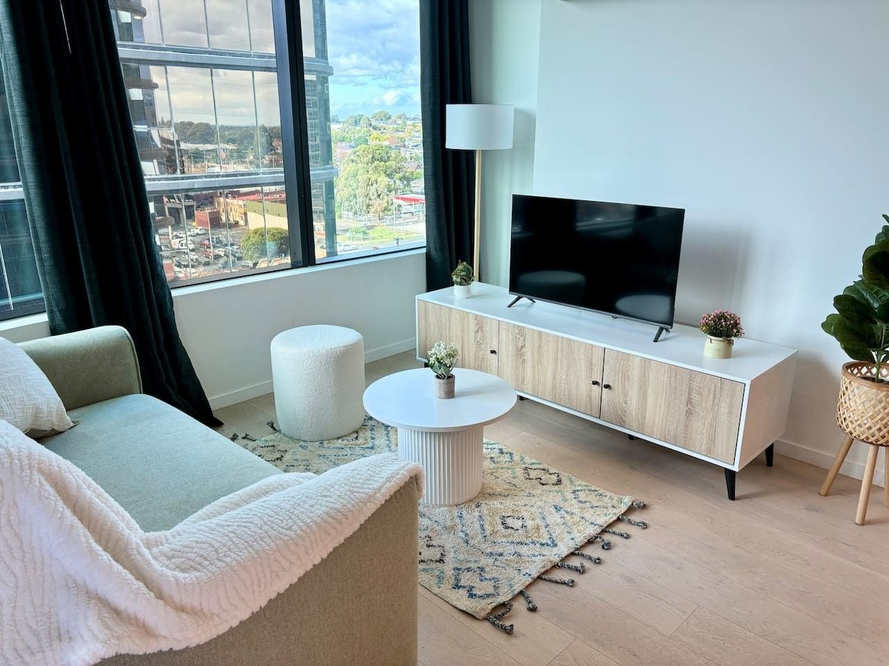 A modern living room with a sofa, a round coffee table, a cabinet with a flat-screen TV, and a large window offering a city view.