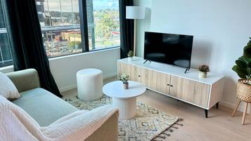 A modern living room with a sofa, a round coffee table, a cabinet with a flat-screen TV, and a large window offering a city view.