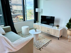 A modern living room with a sofa, a round coffee table, a cabinet with a flat-screen TV, and a large window offering a city view. - SkyGarden Cozy Apt | Sunrise View · Pool · Parking (Glen Waverley)