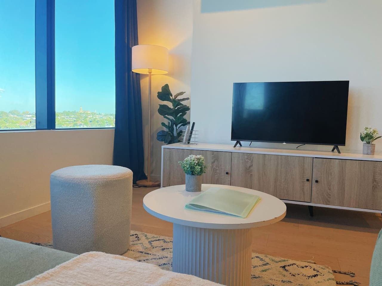 A modern hotel room with a large window, a flat-screen TV, a round white table, a cushioned bench, and a wooden cabinet.