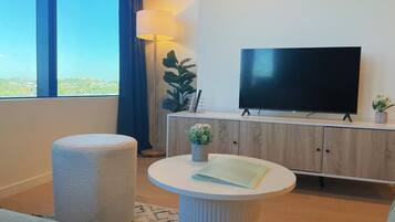 A modern hotel room with a large window, a flat-screen TV, a round white table, a cushioned bench, and a wooden cabinet.