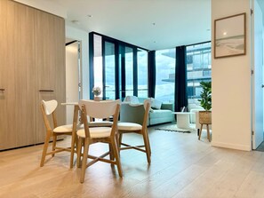 A modern living room with a dining area, large windows, and a view of the outdoors. - SkyGarden Cozy Apt | Sunrise View · Pool · Parking (Glen Waverley)