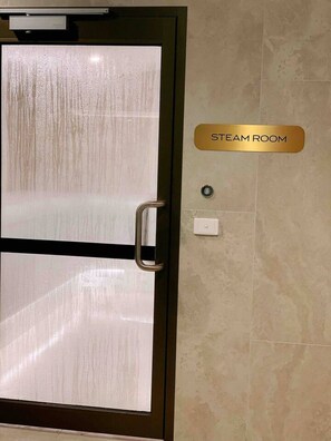 A door with a frosted glass panel and a handle, a sign indicating 'STEAM ROOM', and a light switch on the wall. - SkyGarden Cozy Apt | Sunrise View · Pool · Parking (Glen Waverley)