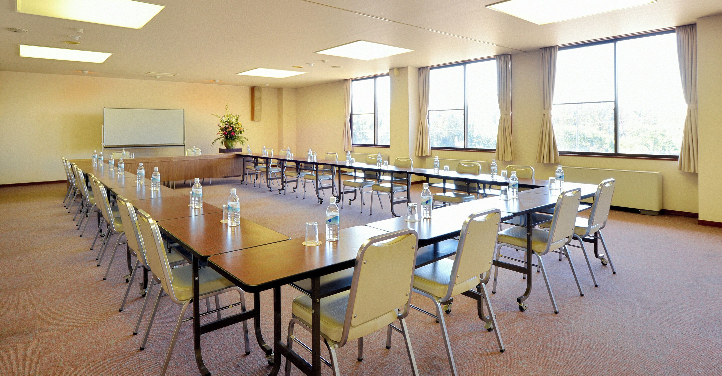 Un salón de conferencias con una larga mesa, sillas, una pizarra y ventanas con cortinas.