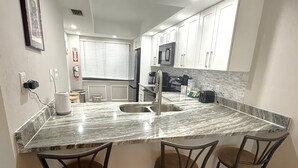 A kitchen with a marble countertop, stainless steel sink, and three bar stools. - Island End Paradise - 2 Bedroom Beachfront (Fort Myers Beach)