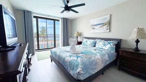 A hotel room with a bed, a desk with a computer, a ceiling fan, a window with a view of the beach, and a painting on the wall. - Island End Paradise - 2 Bedroom Beachfront (Fort Myers Beach)