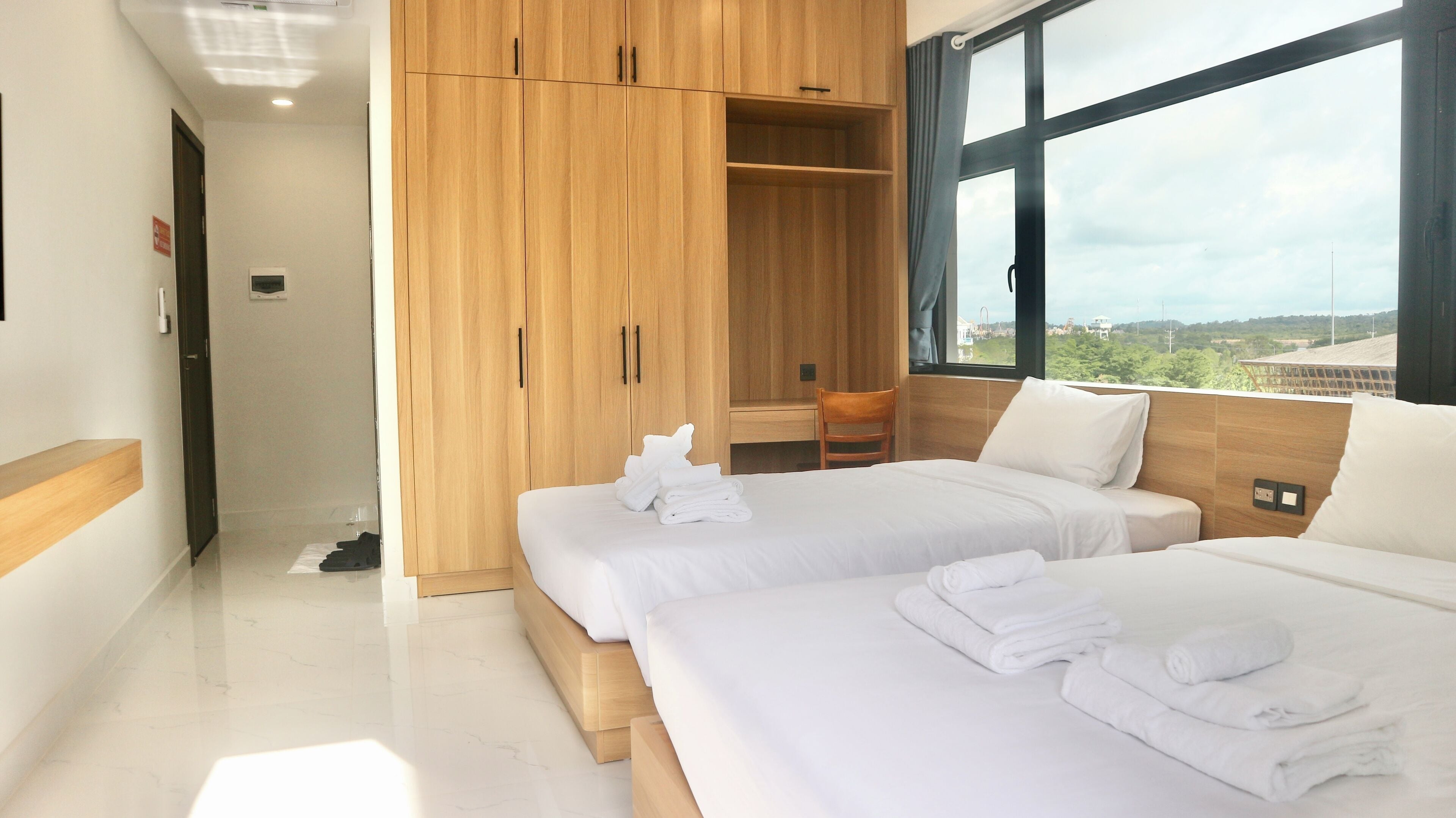A hotel room with two beds, a wooden wardrobe, a desk, and a view of the city.