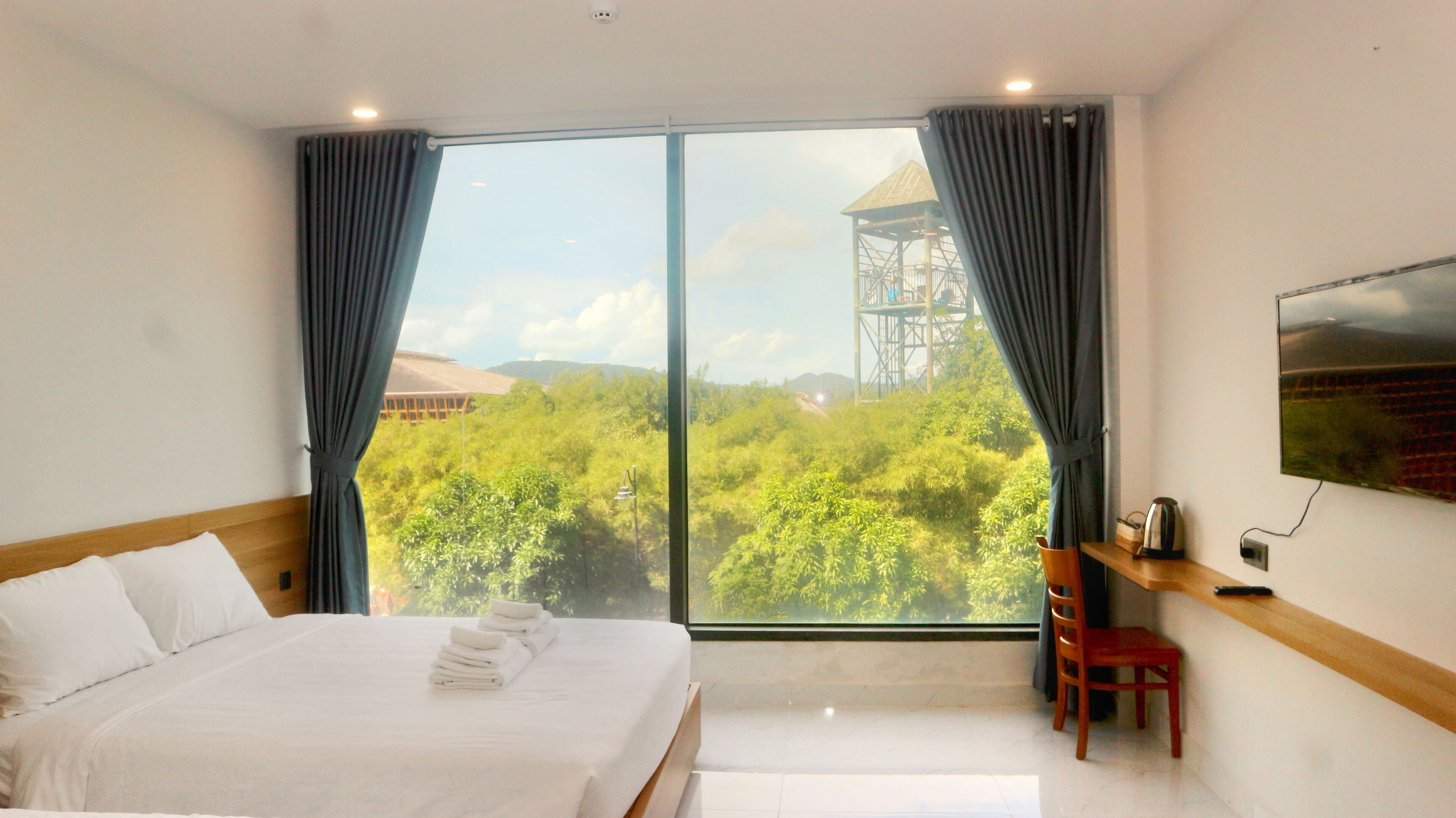 A hotel room with a large bed, a chair, a TV, and a view of a forest and a tower.