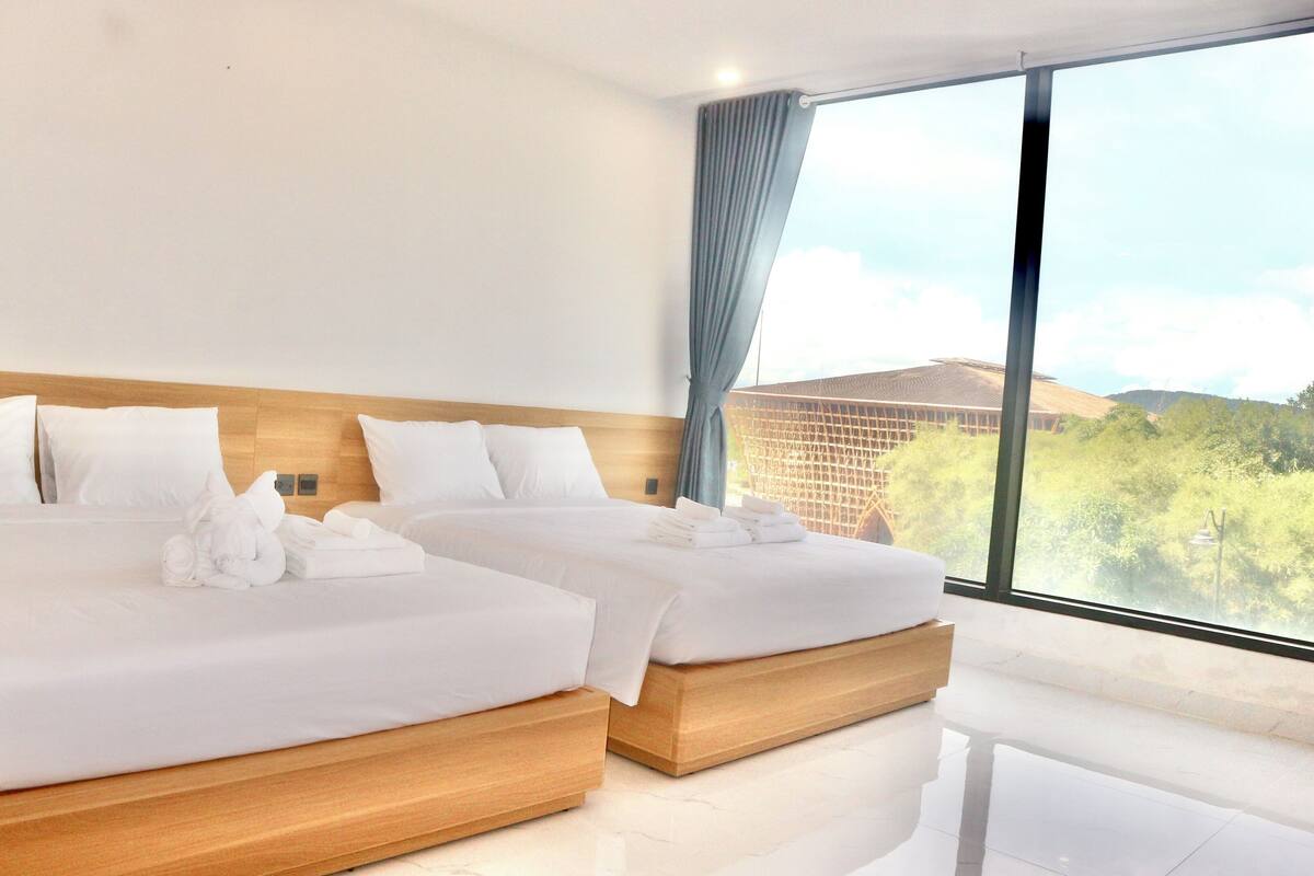 A hotel room with two beds, a wooden headboard, and a large window offering a view of greenery and buildings.