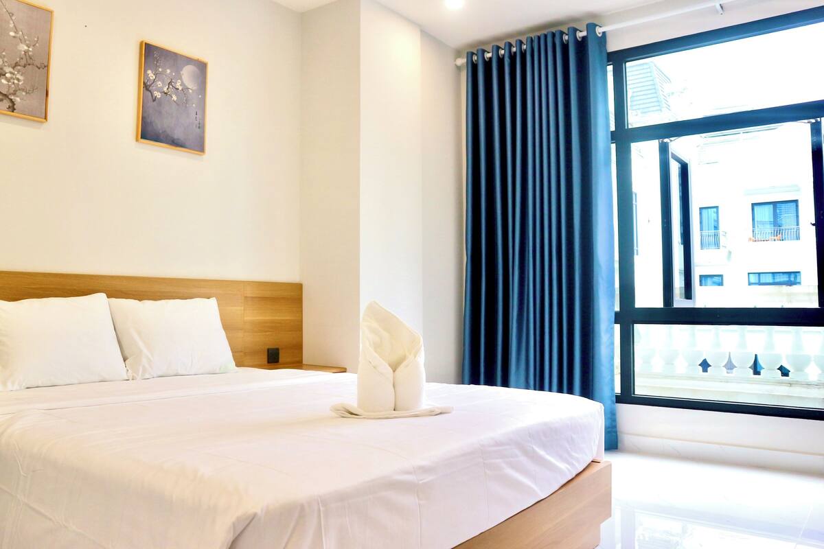 A hotel room with a bed, two pillows, a wooden headboard, a window with curtains, and two wall-mounted artworks.