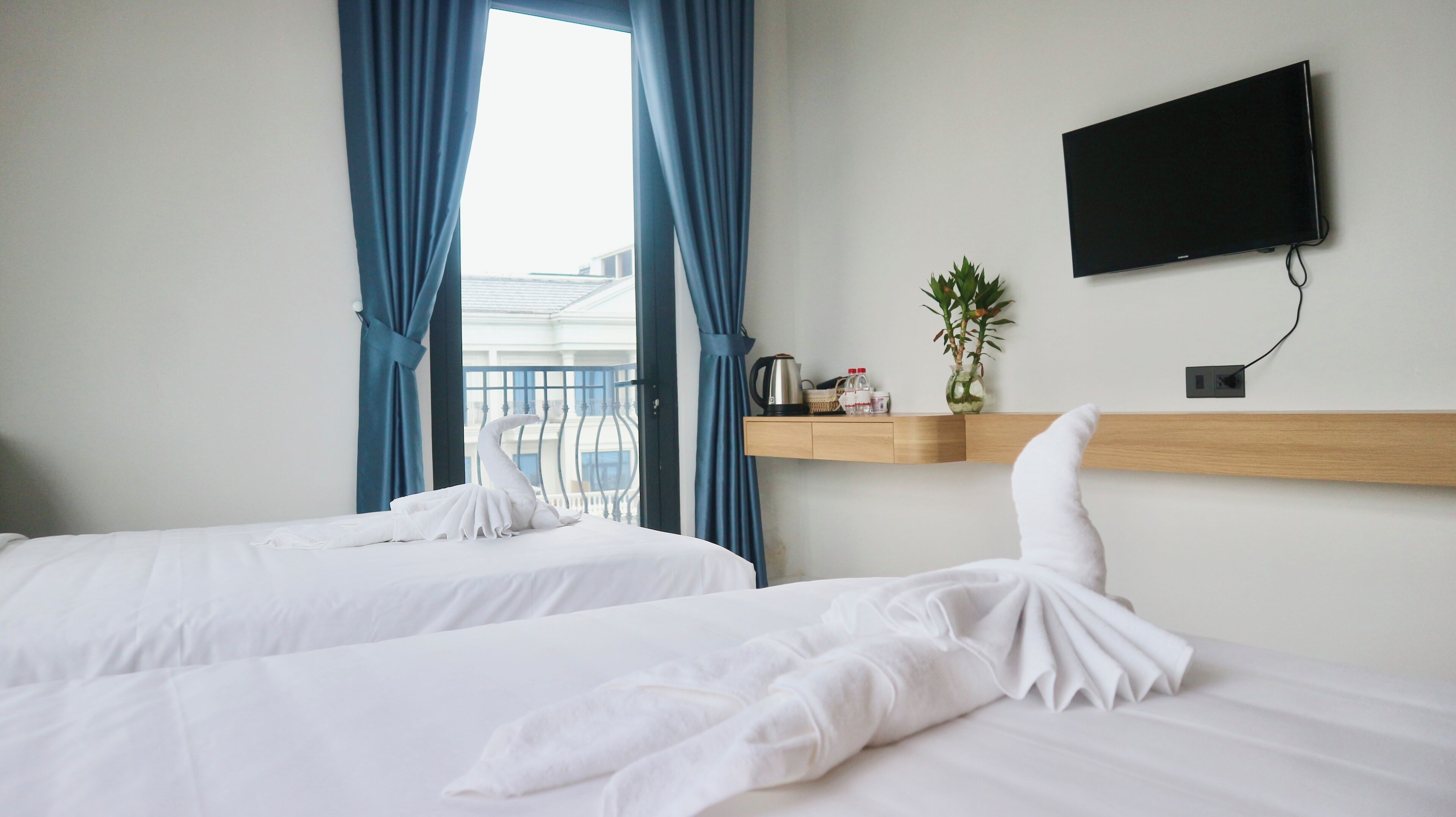 A hotel room with two beds, a TV mounted on the wall, a small shelf with a plant and a kettle, and a view of the outside through large windows.