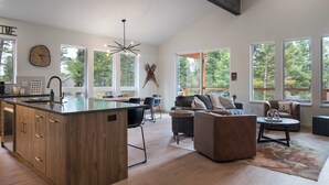 A spacious living area with a large island, modern furniture, and large windows offering views of the outdoors.