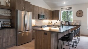 A modern kitchen with wooden cabinets, stainless steel appliances, a central island with bar stools, and a view of the outdoors through a large window.