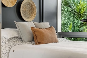 Uma cama com almofadas brancas e castanhas, cabeceira cinzenta e janela com vista para o verde.