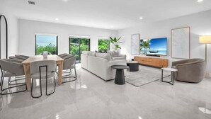 A modern living room with a white sofa, a dining table with chairs, a TV, and a large window with a view of greenery. - Casa Bella l Sleeps 10 l Near Freedom Park & MIA (Miami)