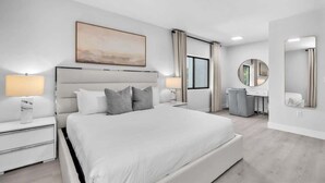 Une chambre d’hôtel moderne avec un grand lit, des tables de chevet avec des lampes, une coiffeuse avec un miroir et un tableau au mur.