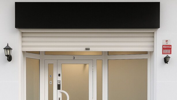 Entrada de hotel com toldo preto, três portas e duas luzes fixadas na parede.