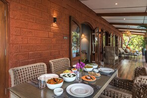 A dining area with a brick wall, wicker chairs, and a table set with various dishes and a vase of flowers. - Amã Stays & Trails Athaang, Dapoli (Dapoli)