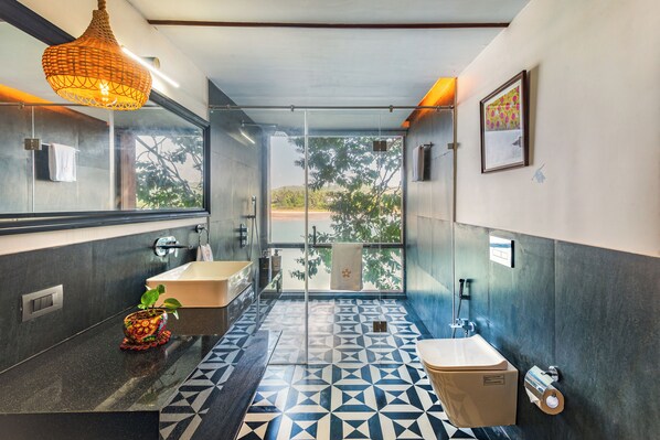 A modern bathroom with a geometric tile floor, a large mirror, a hanging light fixture, and a view of trees through a glass door. - Amã Stays & Trails Athaang, Dapoli (Dapoli)