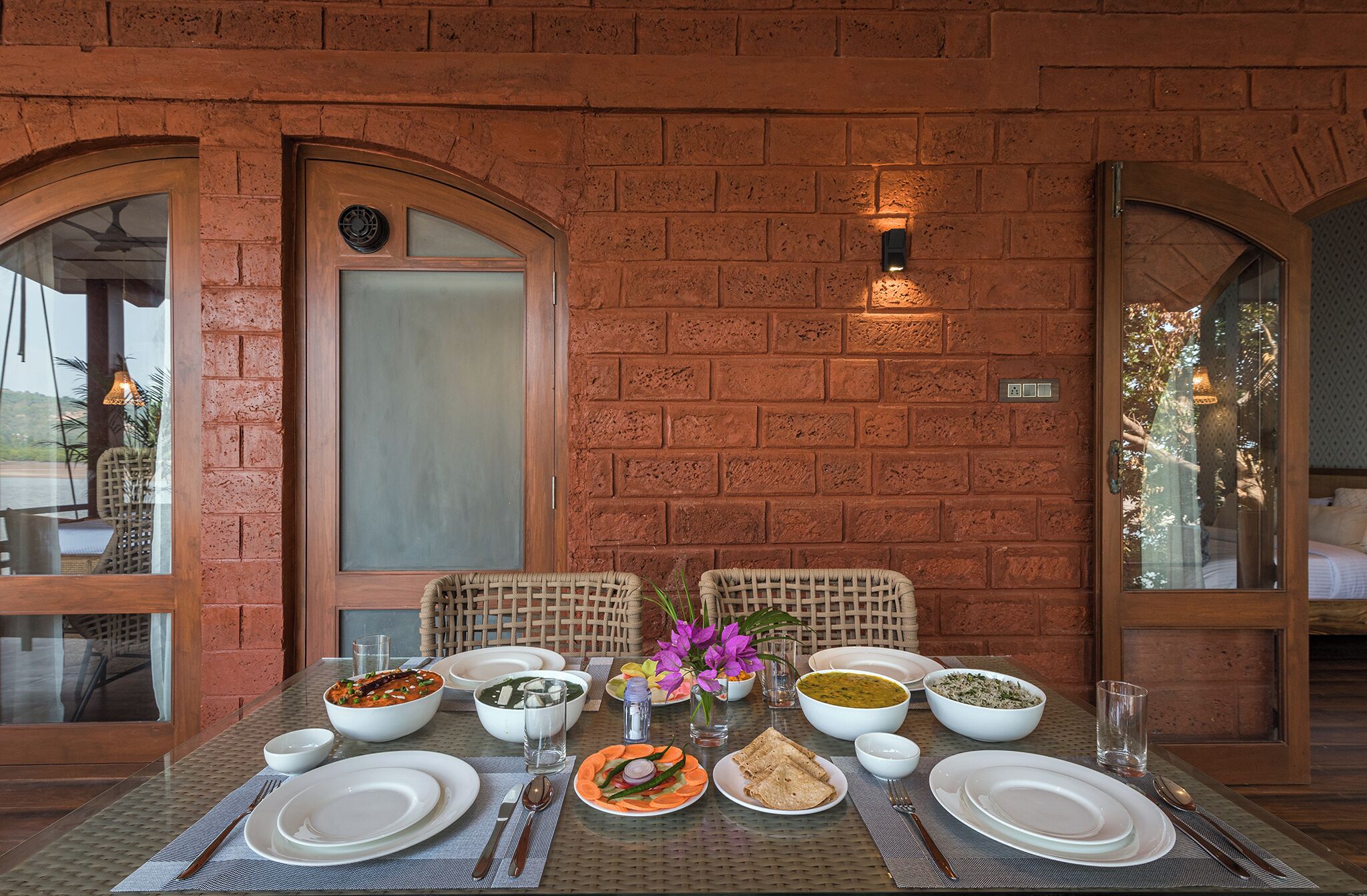 A dining table set with various dishes, surrounded by wicker chairs, against a brick wall with arched windows.
