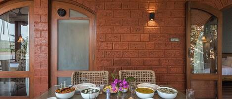 A dining table set with various dishes, surrounded by wicker chairs, against a brick wall with arched windows.